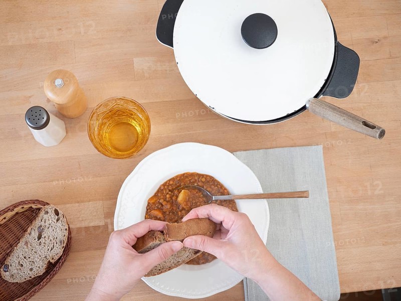 Hands breaking slice of bread over bowl of lentil soup, served with drink, salt and pepper, and bread basket, top view