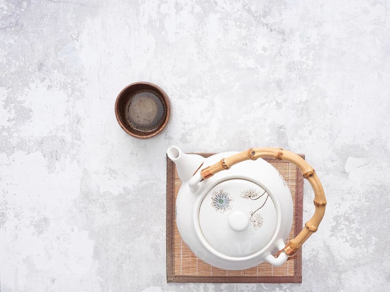 White teapot with bamboo handle and small wooden teacup on bamboo mat, ready for traditional asian tea ceremony