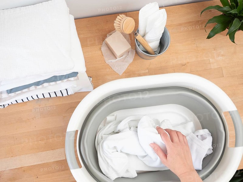 Hand soaking white clothes in basin with natural soap and brush on wooden table, promoting sustainable laundry practices