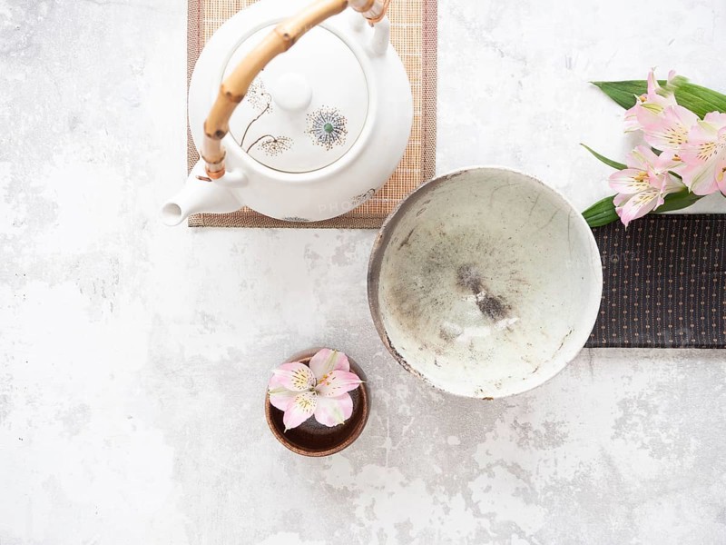 Asian tea ceremony arrangement with white teapot, empty bowl, pink flowers and bamboo placemat on white background