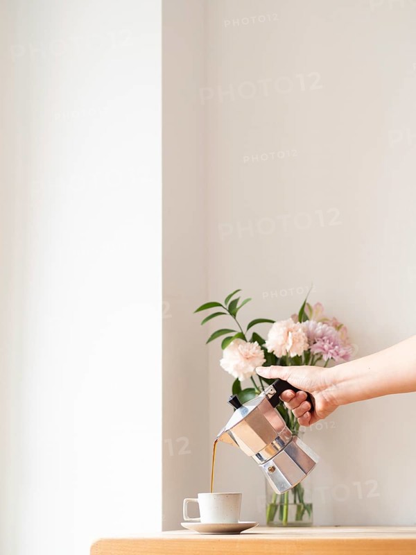 Hand pouring coffee from moka pot into cup on wooden table, with flowers in background, in minimalist cafe setting