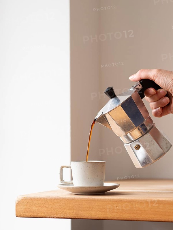 Hand pouring coffee from a moka pot into a cup, placed on a wooden table, creating a cozy and aromatic scene