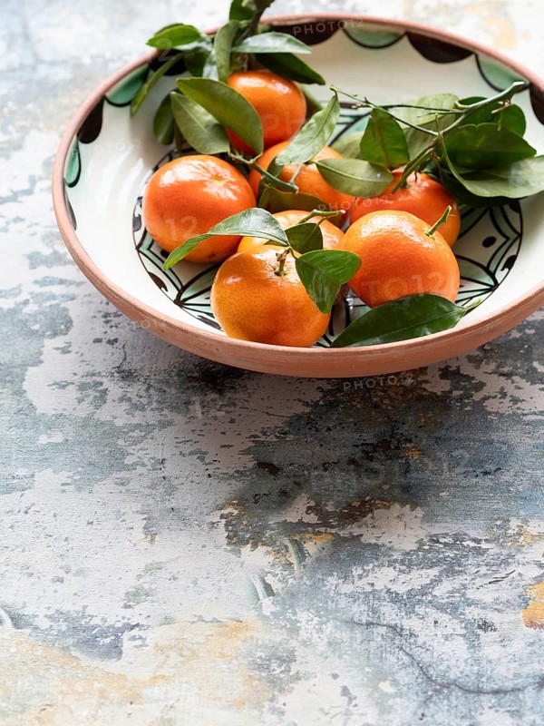 Vibrant tangerines with green leaves arranged in a decorative ceramic bowl, set against a rustic tabletop, creating a still life composition