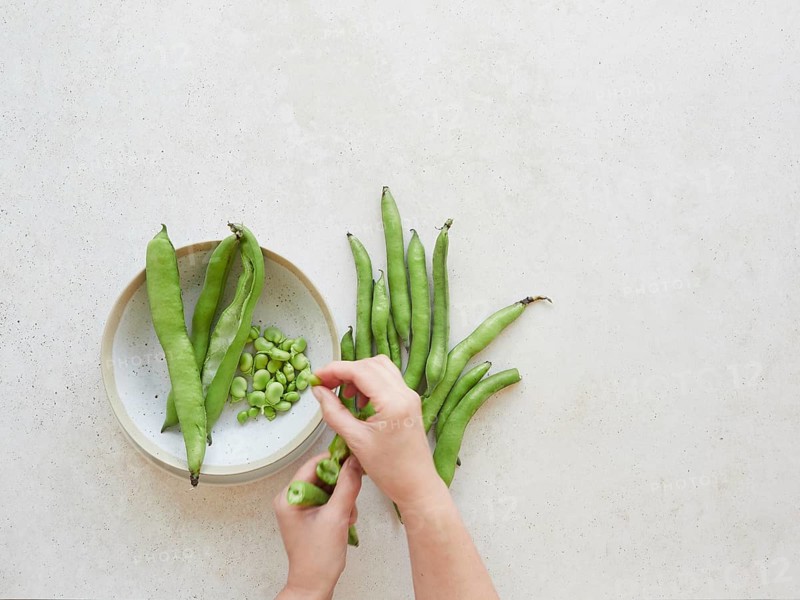 Hands shelling fresh broad beans, also known as fava beans, on a kitchen counter, ready for a healthy and delicious vegetarian meal