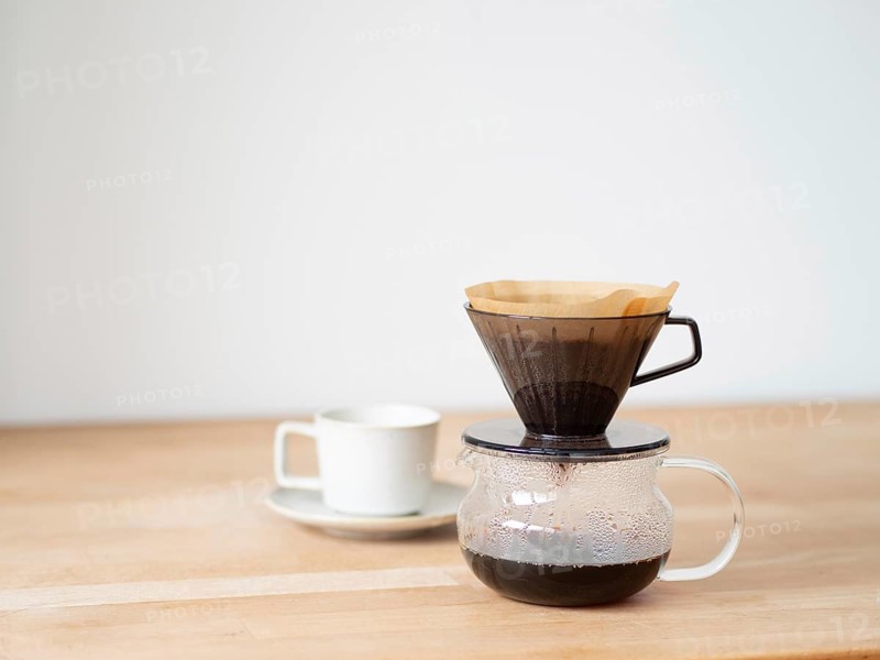 Coffee is brewing using a paper filter and dripper on a wooden table, with a white cup and saucer nearby