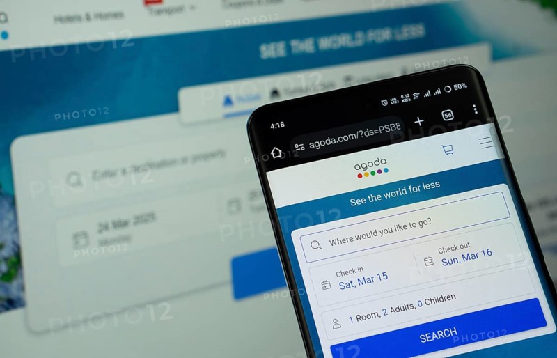 Dhaka, Bangladesh - 27 July 2025: The logo of Agoda is displayed on a smartphone screen, and the website is visible on a computer screen in the background