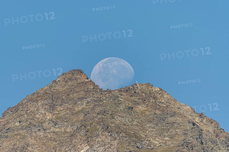 Majestic mountain peak stands tall against vibrant blue sky as large moon rises just behind it. The scene captures the quiet beauty of nature in the early evening light. Grachen, Viege, Valais, Switzerland