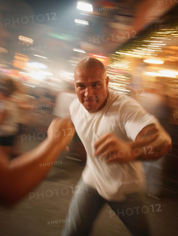 A man gets into a fighting stance, surrounded by dynamic movement and a background filled with lights, symbolic image for physical confrontation in public, use of force, physical violence, act of violence, aggressive behaviour, brawl, AI generated