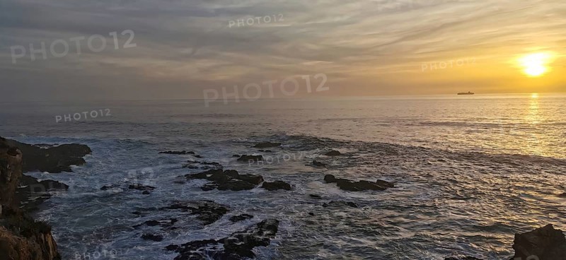The picture shows a sunset over the sea with rocks in the foreground, hiking on the Fisherman's trail, Rota Vicentina, Porto Covo, Alentejo region, Portugal