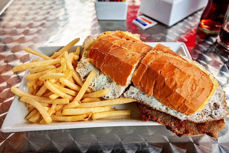 Large sandwich with fries served on a metal table, thickly filled with bread and a fried egg visible, An Argentinean Lomito