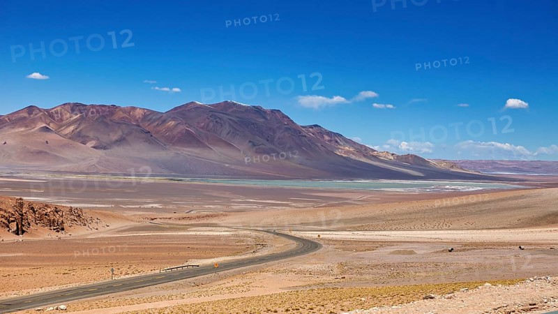 A long road snakes through an arid desert landscape with distant mountains, the countryside and colorful rock formations of the Quebrada de Cafayate near Salta in Argentina