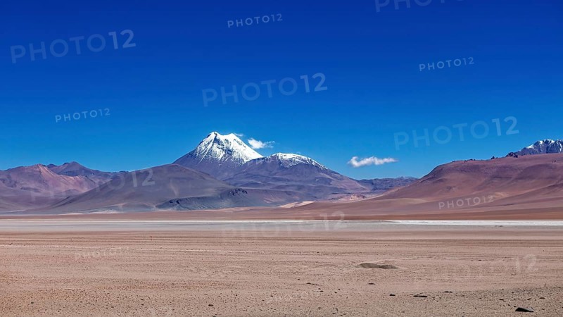 A snow-covered volcano rises above a dry, vast desert under clear skies, the landscape and colorful rock formations of the Quebrada de Cafayate near Salta in Argentina