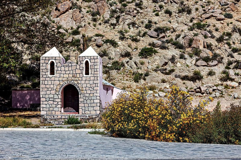 Small stone house in a desert area with yellow flowers in the foreground against a rocky mountain backdrop, The chapel in Santa Rosa de Tastil near Salta in Argentina
