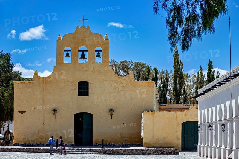 Yellow church with bell tower under blue sky, people on stairs surrounded by trees, Iglesia San José de Cachi in Argentina