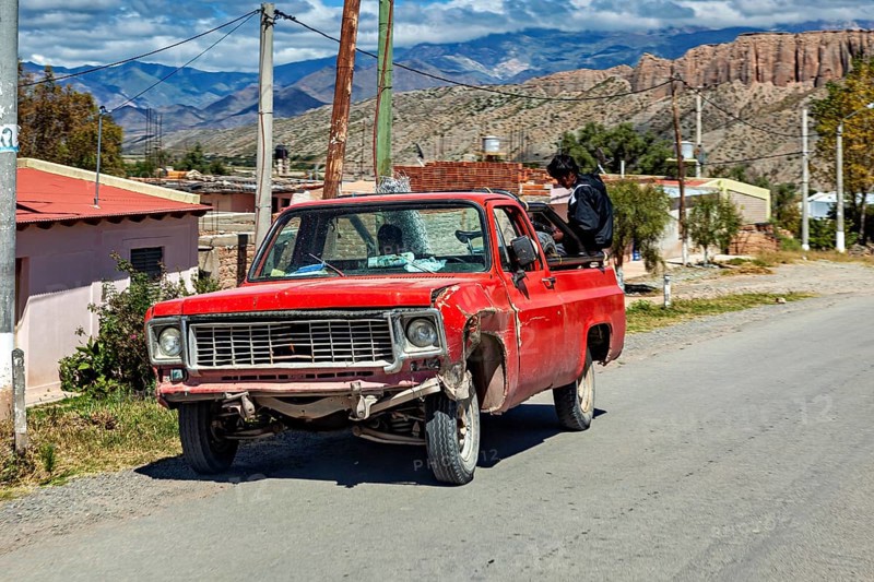 Red, slightly damaged pickup truck on a country road with a man and mountain scenery, old cars in Argentina