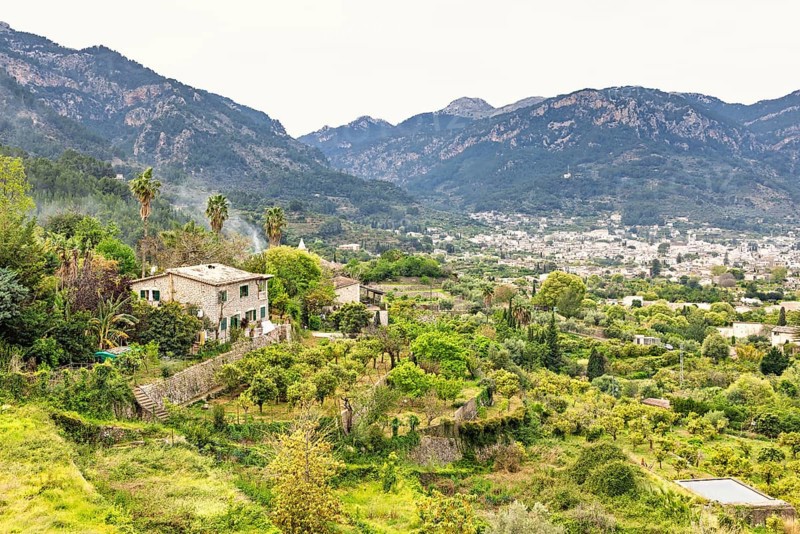 Serene rural landscape featuring a traditional stone house surrounded by vibrant greenery and terraced gardens with a city in the mountain valley, Sóller, Mallorca, Spain