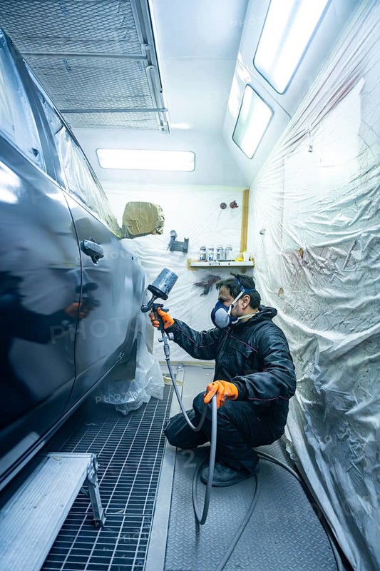 Automotive technician wearing protective mask and gloves, using a spray gun to apply fresh paint to a vehicle in a modern, well lit spray booth, carefully repairing the car body