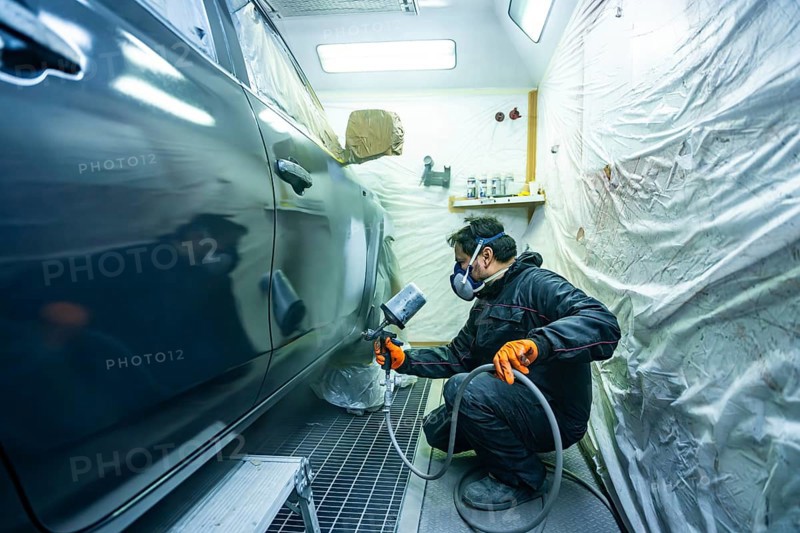 Mechanic wearing respirator mask and orange gloves spraying dark car in a professional paint booth, performing auto body repair and re painting services