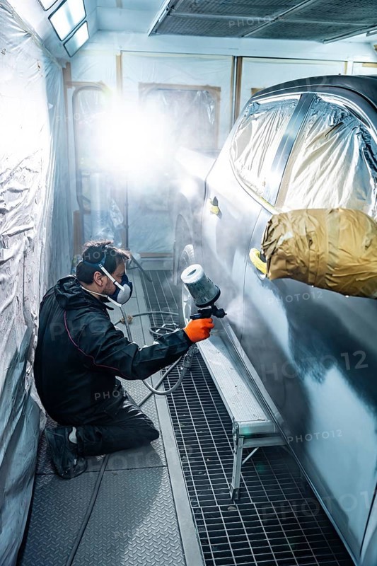 Professional auto body mechanic kneeling while applying primer or paint to a passenger cars side door using a spray gun in a clean, brightly lit paint booth