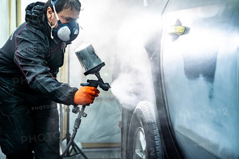 Skilled automotive technician in protective gear applying fresh paint to a car door using a spray gun, performing surface conditioning work in a dedicated car paint booth