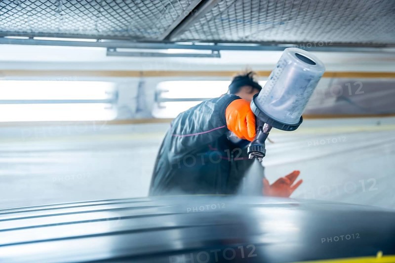Auto body technician wearing protective gloves painting a vehicle car roof using a spray gun in a well lit automotive service workshop, ensuring high quality refinishing and repair work