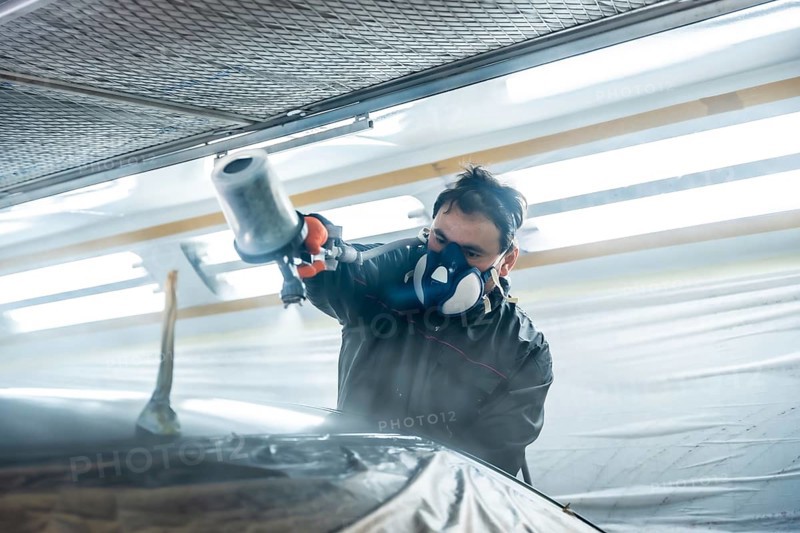 Auto body technician wearing protective respirator and suit painting a car part with a spray gun, performing professional repair work in a vehicle paint booth