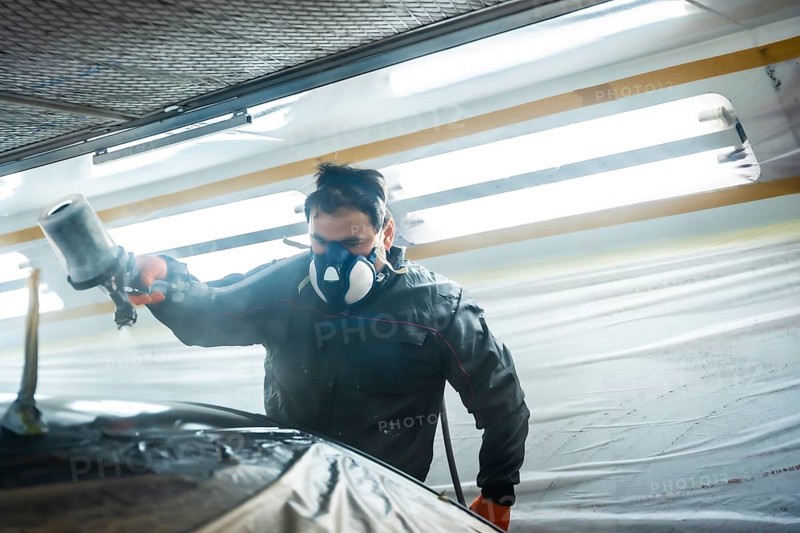 Auto body technician working in a specialized workshop, wearing a respirator mask and protective suit while applying fresh paint to a car part with a paint gun