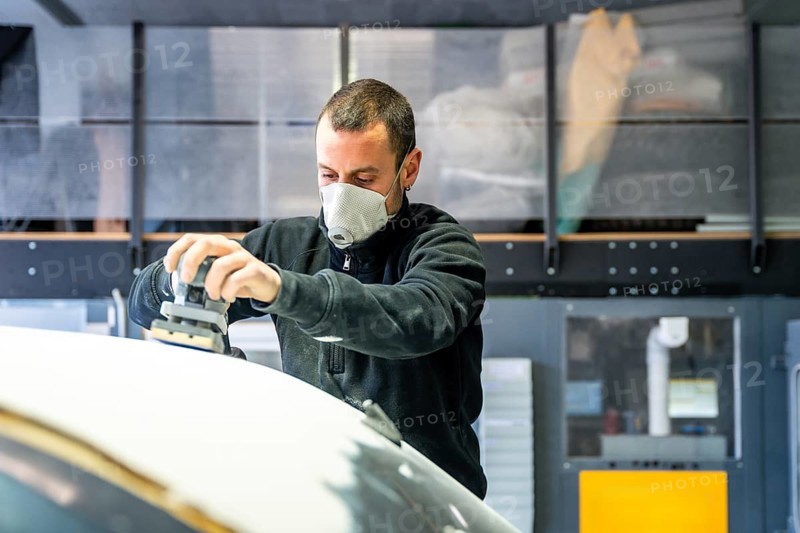 Auto body technician wearing a respirator mask using an orbital sander to prepare a car surface for painting, managing dust in a professional garage environment