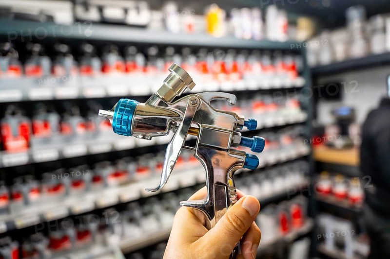 Hand holding a high pressure spray gun, selecting paint colors from shelves in an automotive refinishing workshop, preparing for car body repair and painting