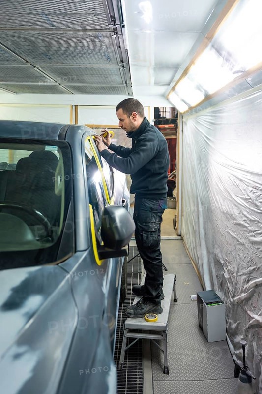 Mechanic applying masking tape meticulously on a car body before painting in a professional automotive repair and service workshop, ensuring precise preparation for a flawless finish