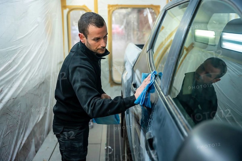 Man carefully wiping car body panels with a blue microfiber cloth inside a professional spray booth, preparing the vehicle for painting precise automotive detailing and repair work