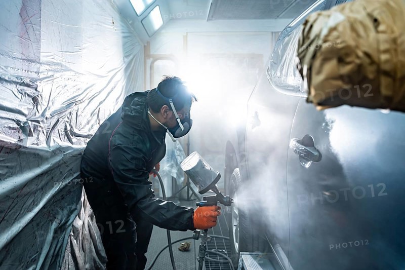 Professional automotive technician applying a fresh coat of paint to a vehicle in a specialized spray booth, wearing a respirator mask and protective gear during the careful repainting process