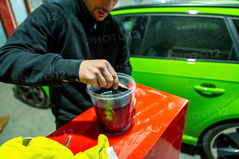 Male worker preparing automotive paint in a plastic container with a lime green vehicle visible in the background, focusing on the careful process of color matching and repair
