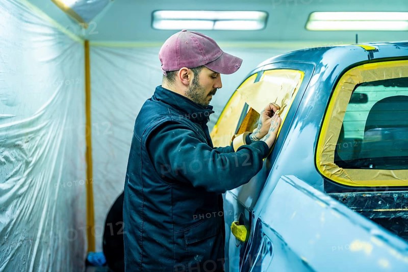 Auto body technician carefully applying masking tape and paper to a car window, preparing the vehicle for a new paint job inside a professional automotive repair paint booth