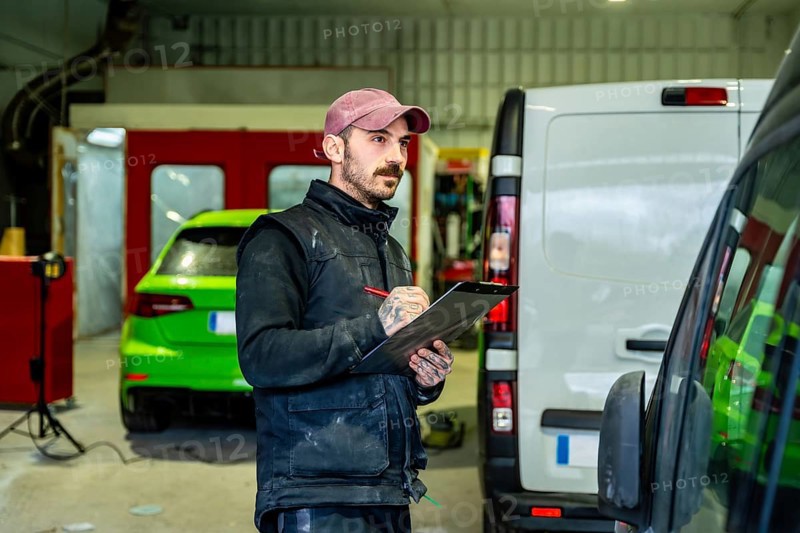 Auto body shop technician checking details on a white van and a green car in the background, working with a clipboard and pen in a professional automotive service garage