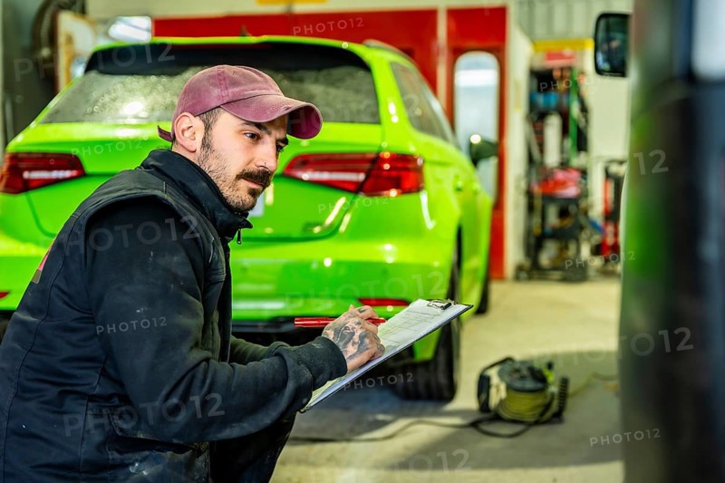 Automotive technician crouching and writing notes on a clipboard during a vehicle inspection or repair assessment at a professional car body and paint shop