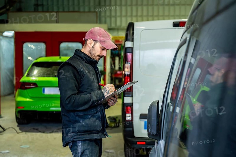 Auto body technician wearing a cap and vest carefully inspecting a vehicle and writing notes on a clipboard, standing between a white van and a neon green car in a repair garage