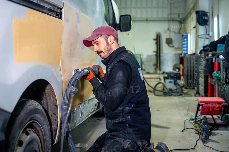Auto body technician working on a van, utilizing a power sander with vacuum attachment to smooth filler on the vehicle's side panel at an automotive repair workshop