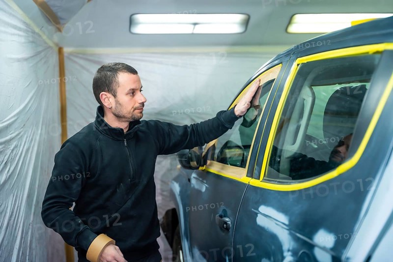 Auto body shop worker carefully masking a vehicle's windows and trim with tape, preparing the surface for a professional paint job after repairs in a garage