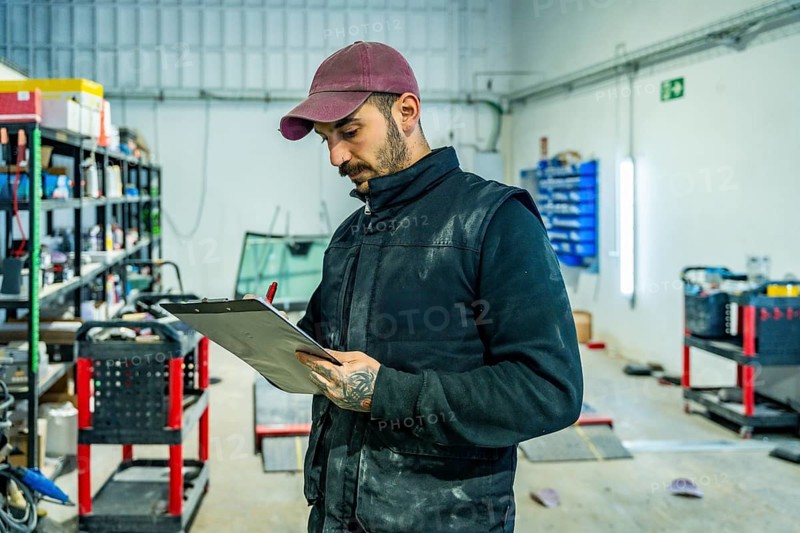 Male technician wearing a cap and vest standing in an automotive workshop, holding a clipboard and pen, reviewing a checklist for vehicle car body repairs or maintenance
