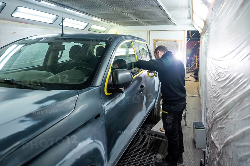 Automotive technician preparing a vehicle for painting by carefully applying protective masking tape around windows and trim in a bright auto body repair booth