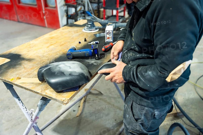 Auto body repairman meticulously sanding a car part, preparing surfaces for paint spraying, restoring vehicle bodywork in a professional automotive workshop with tools on a wooden table