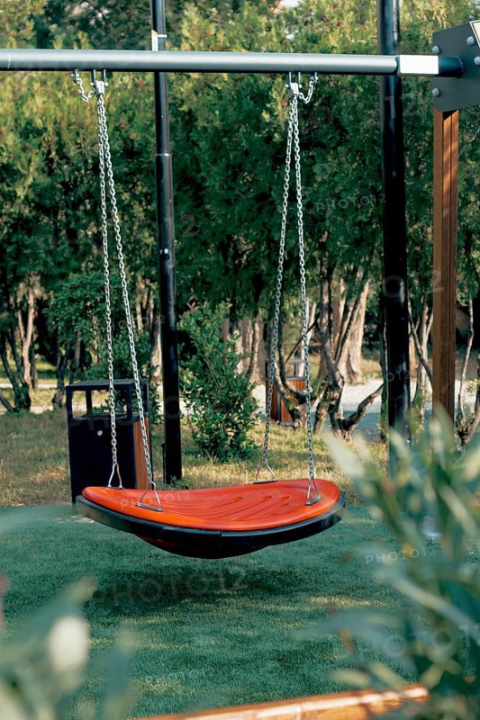 A bright orange hanging swing is set in a serene garden surrounded by trees and grass. The peaceful environment invites relaxation and enjoyment on a sunny afternoon