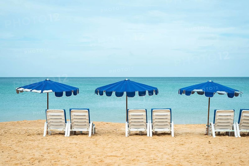 A serene beach scene with three blue umbrellas and white lounge chairs on golden sand facing a calm, expansive ocean in Thailand. The tranquil setting invites relaxation and leisure in vacation
