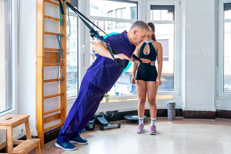 Physician assisting male patient performing strength exercises with suspension straps, guided by a female trainer in a modern physiotherapy and rehabilitation clinic using functional training