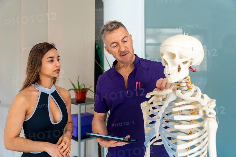 Experienced osteopath or physiotherapist showing a female patient spinal anatomy on a human skeleton model during a medical recovery or rehabilitation consultation in a clinic