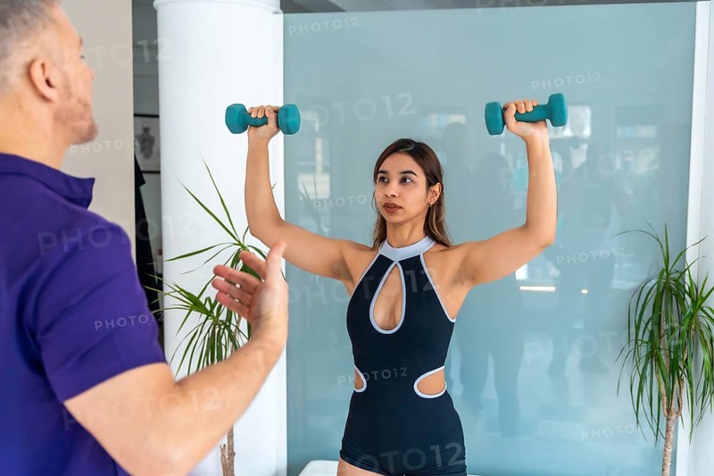 Physiotherapist instructing a focused young woman performing light dumbbell exercises as part of her rehabilitation and medical recovery program in a bright, modern clinic
