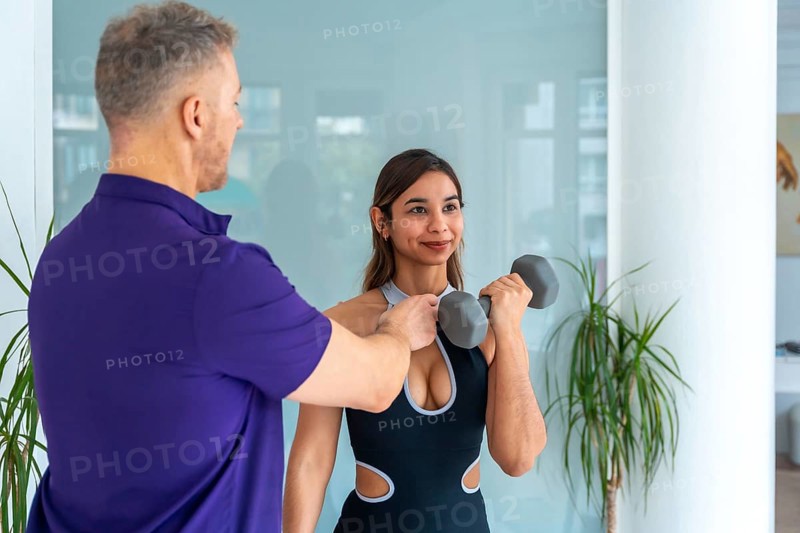 Physiotherapist male trainer assisting a young woman client with rehabilitation and strength training exercises using a dumbbell, focusing on medical recovery and physical therapy in a gym setting