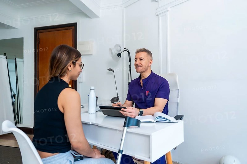 Healthcare professional wearing a uniform discussing medical recovery and sports osteopathy with a young female patient in a clinic office, developing a customized physiotherapy treatment