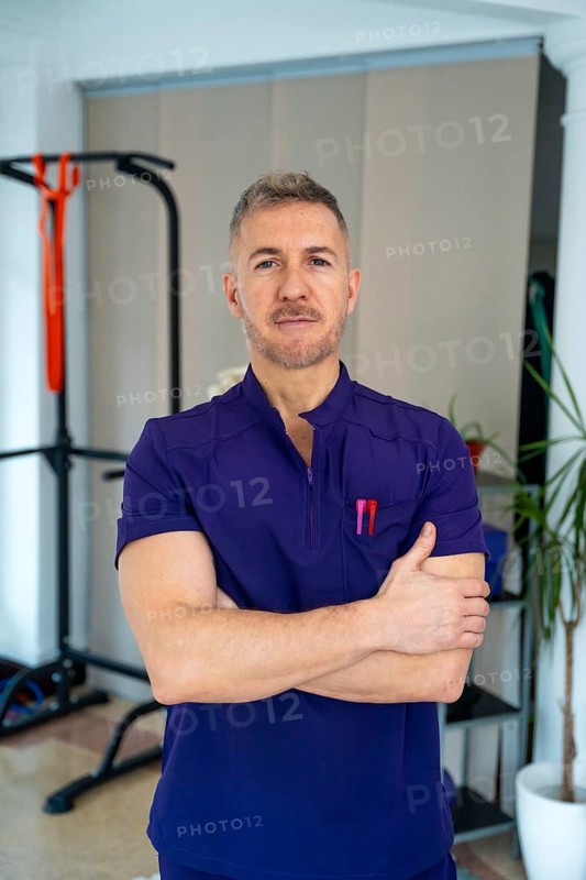 Male physiotherapist in purple uniform stands with arms crossed in a clinic, representing concepts of sports medicine, rehabilitation, and medical recovery